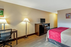 Quarto de hotel com cama, escrivaninha, cadeira, luminária, televisão e um pequeno armário.