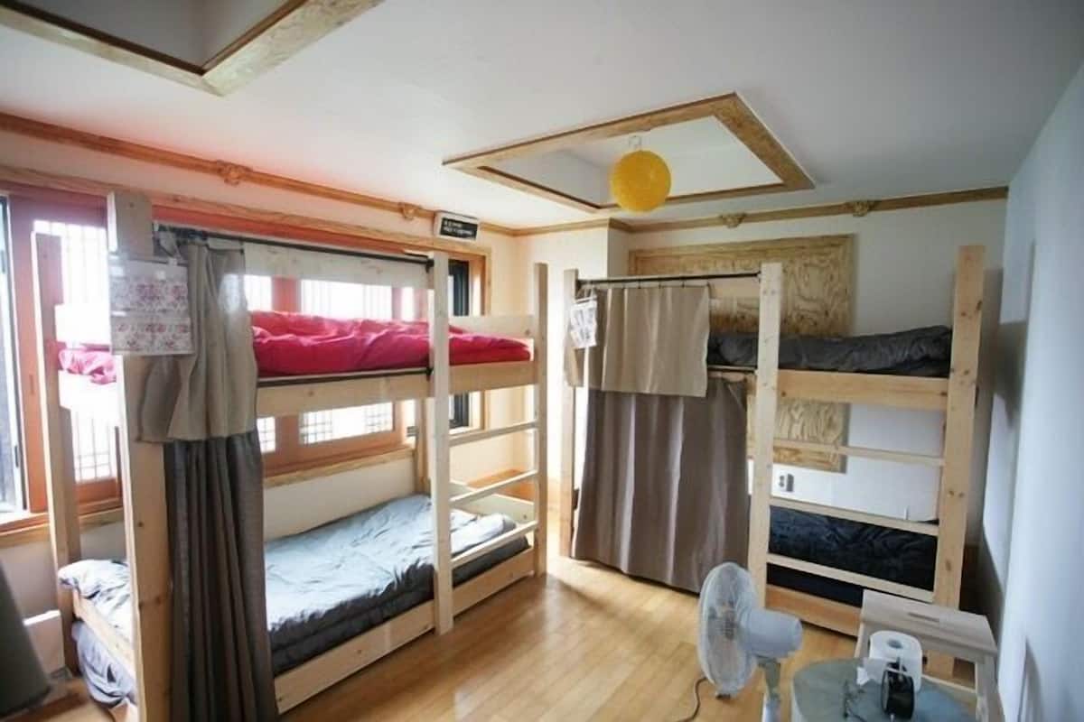 Une chambre de dortoir avec des lits superposés, un ventilateur et une petite table avec une lampe.