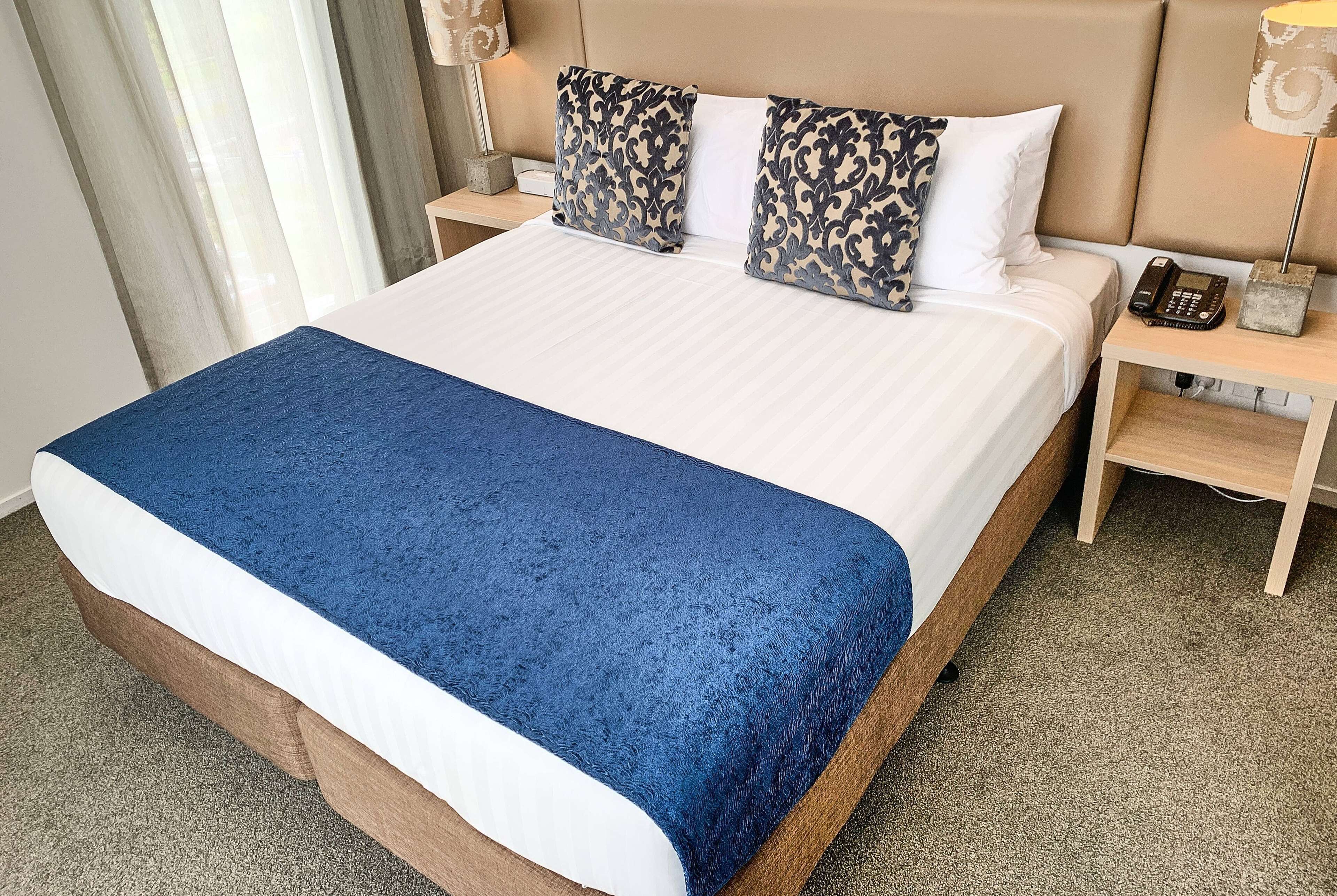 A neatly made bed with a blue blanket, two patterned pillows, and a bedside phone on a wooden nightstand.