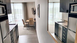 Una cocina moderna con armarios oscuros, un comedor con mesa y sillas, y vistas a través de una ventana con cortinas translúcidas.