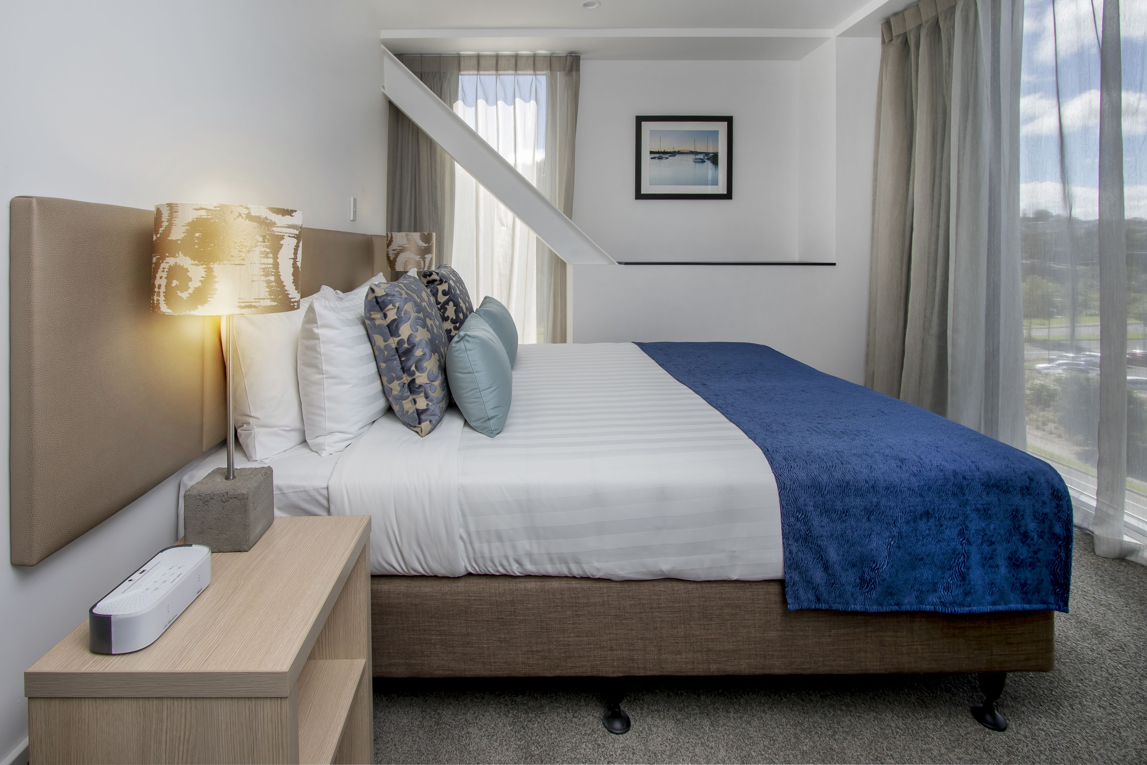 A hotel room with a bed, a nightstand with a lamp, a remote control, a picture on the wall, and a view of the outside.