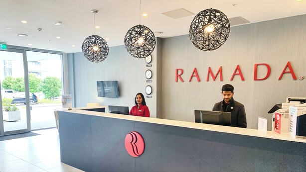Una recepción con dos empleados, un gran letrero de 'RAMADA' y un diseño interior moderno con lámparas colgantes esféricas.