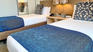 Una habitación de hotel con dos camas, cada una con una manta azul estampada, una mesita de noche con lámpara y un espejo en la pared.
