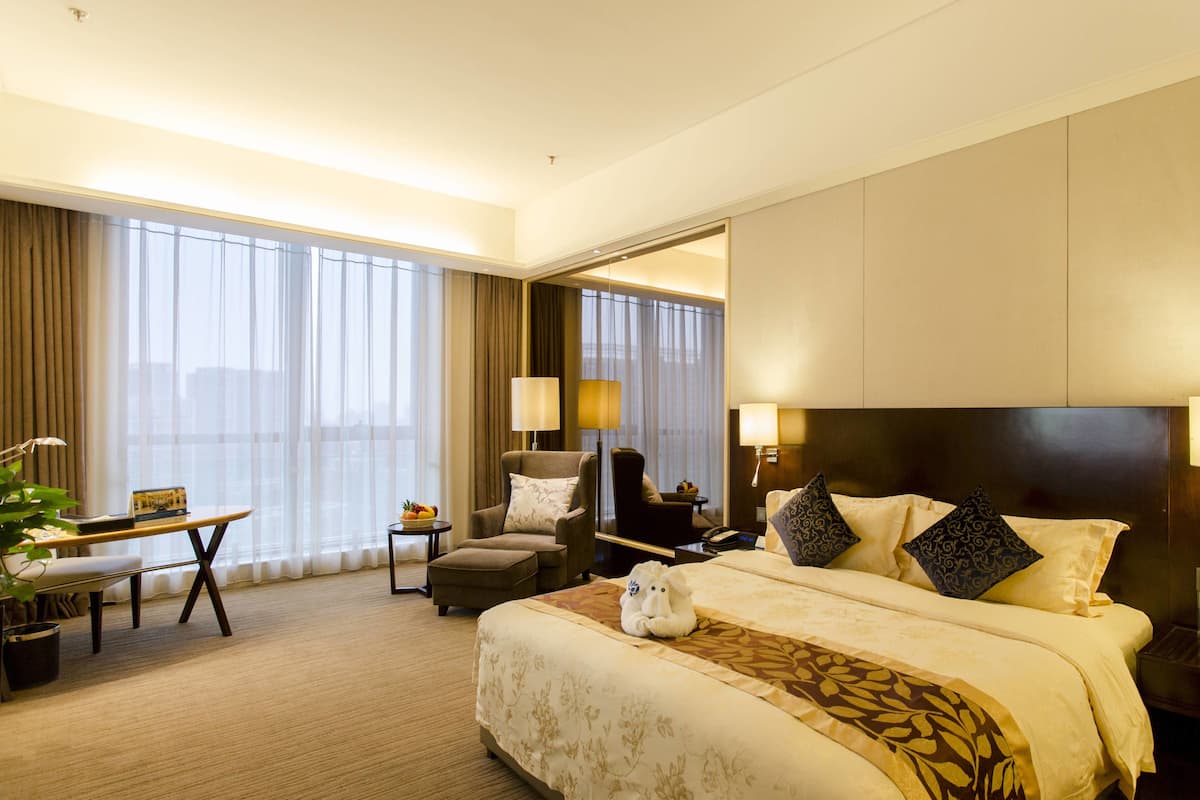 Una habitación de hotel con una cama grande, un escritorio con una silla, una mesita con frutero y un ventanal con cortinas.