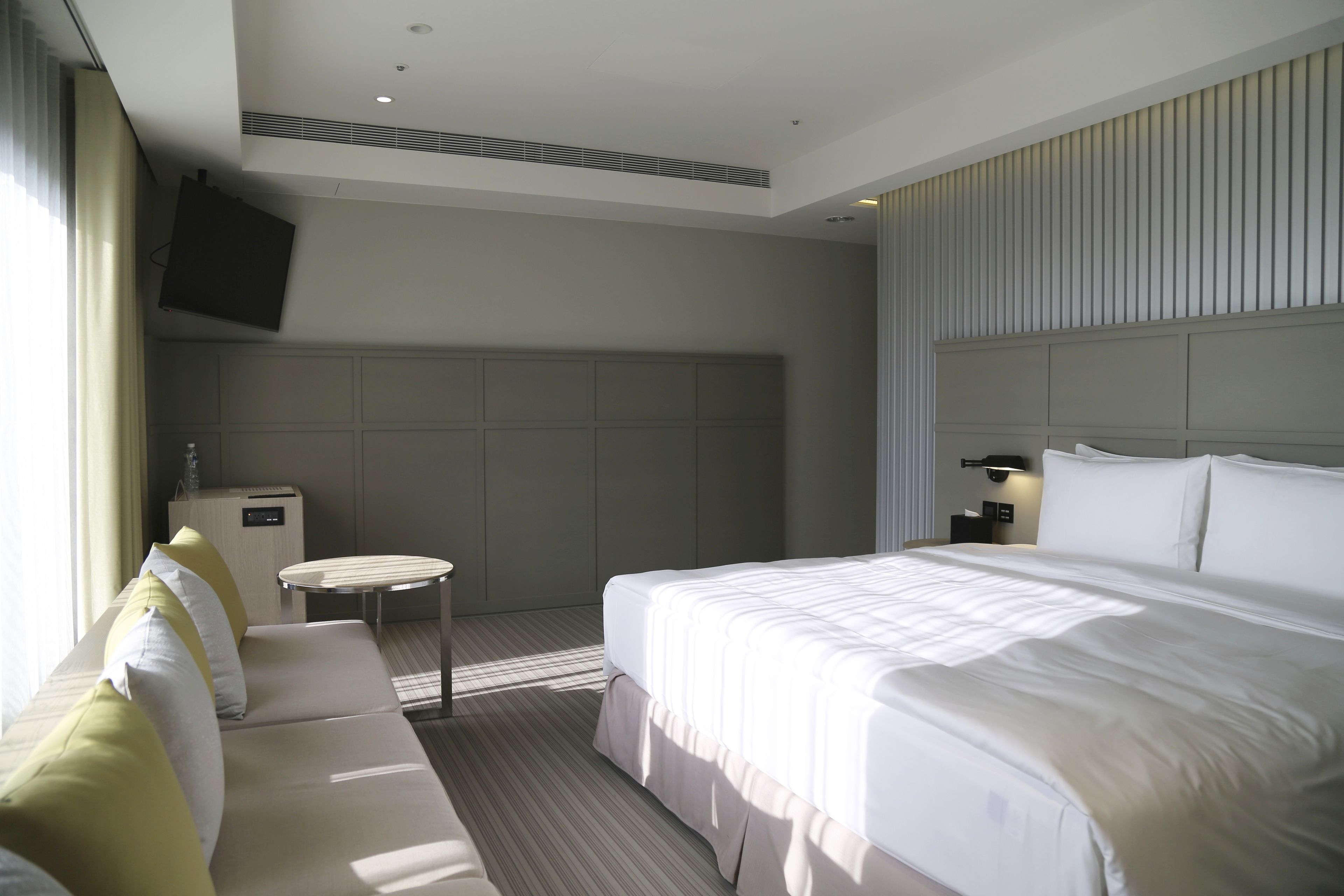 A hotel room with a large bed, a sofa, a small table, a TV mounted on the wall, and a control panel.