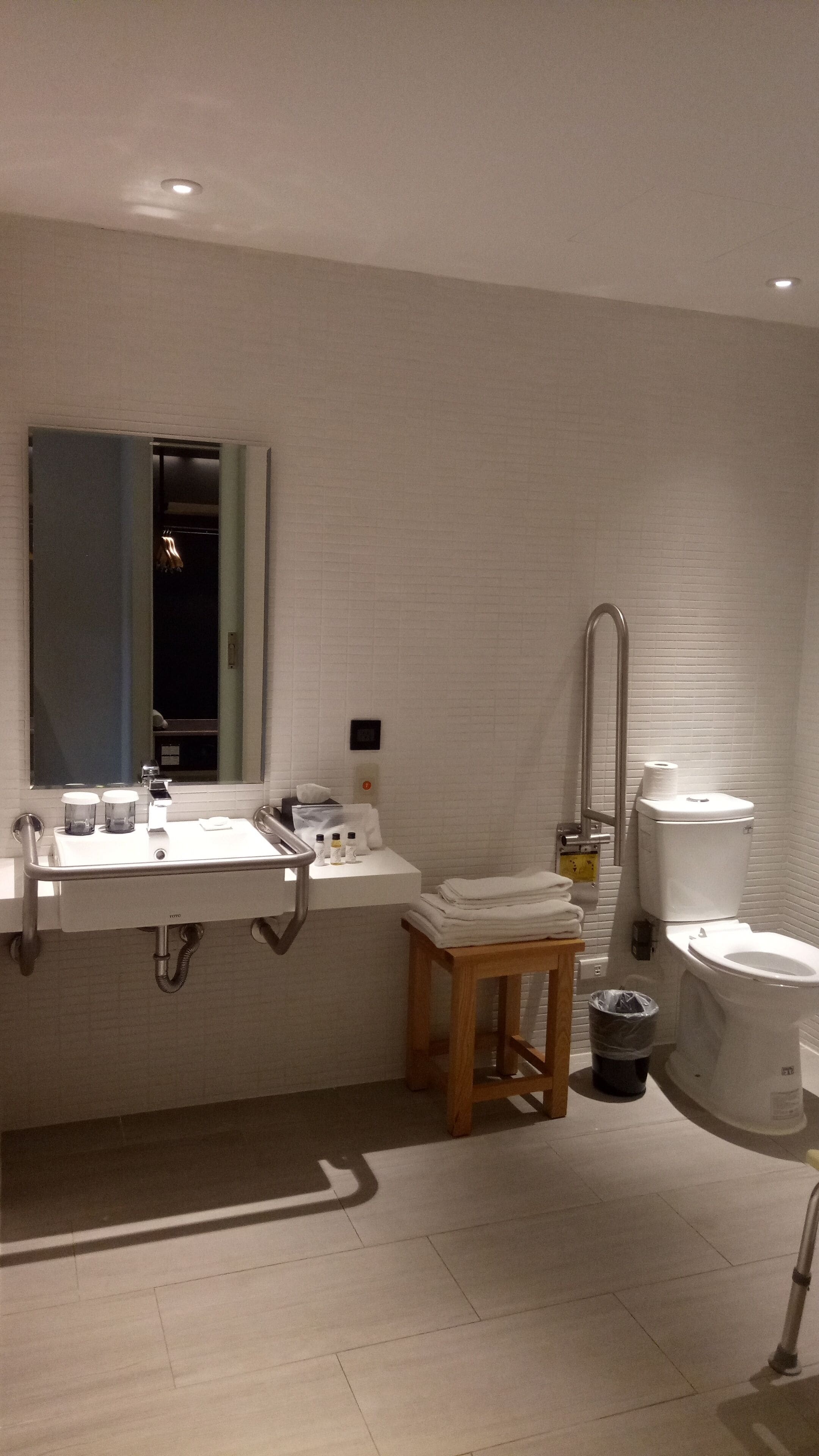 A modern bathroom with a white sink, mirror, and toilet. There is a wooden stool and a trash bin.