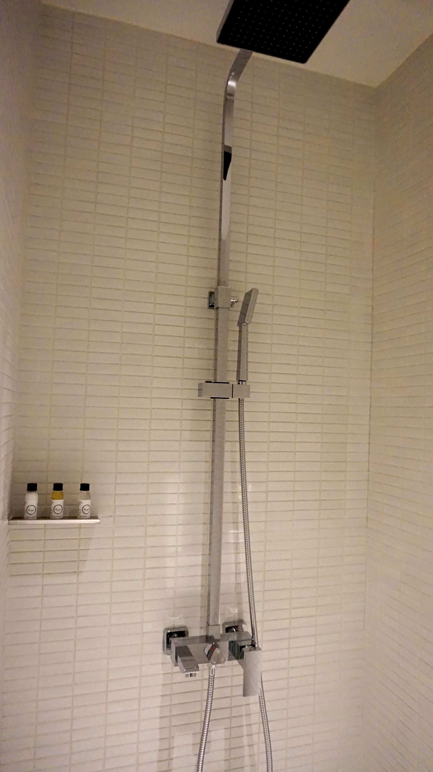 A shower with a handheld showerhead and a fixed showerhead, both mounted on a wall with a tiled backsplash. There is a shelf with three bottles to the left of the shower.