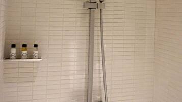 A shower with a handheld showerhead and a fixed showerhead, both mounted on a wall with a tiled backsplash. There is a shelf with three bottles to the left of the shower.