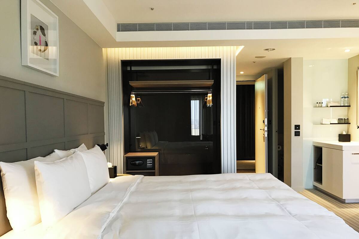 A modern hotel room with a large bed, a bedside table, a mirror, and a bathroom area with a sink and shelves.