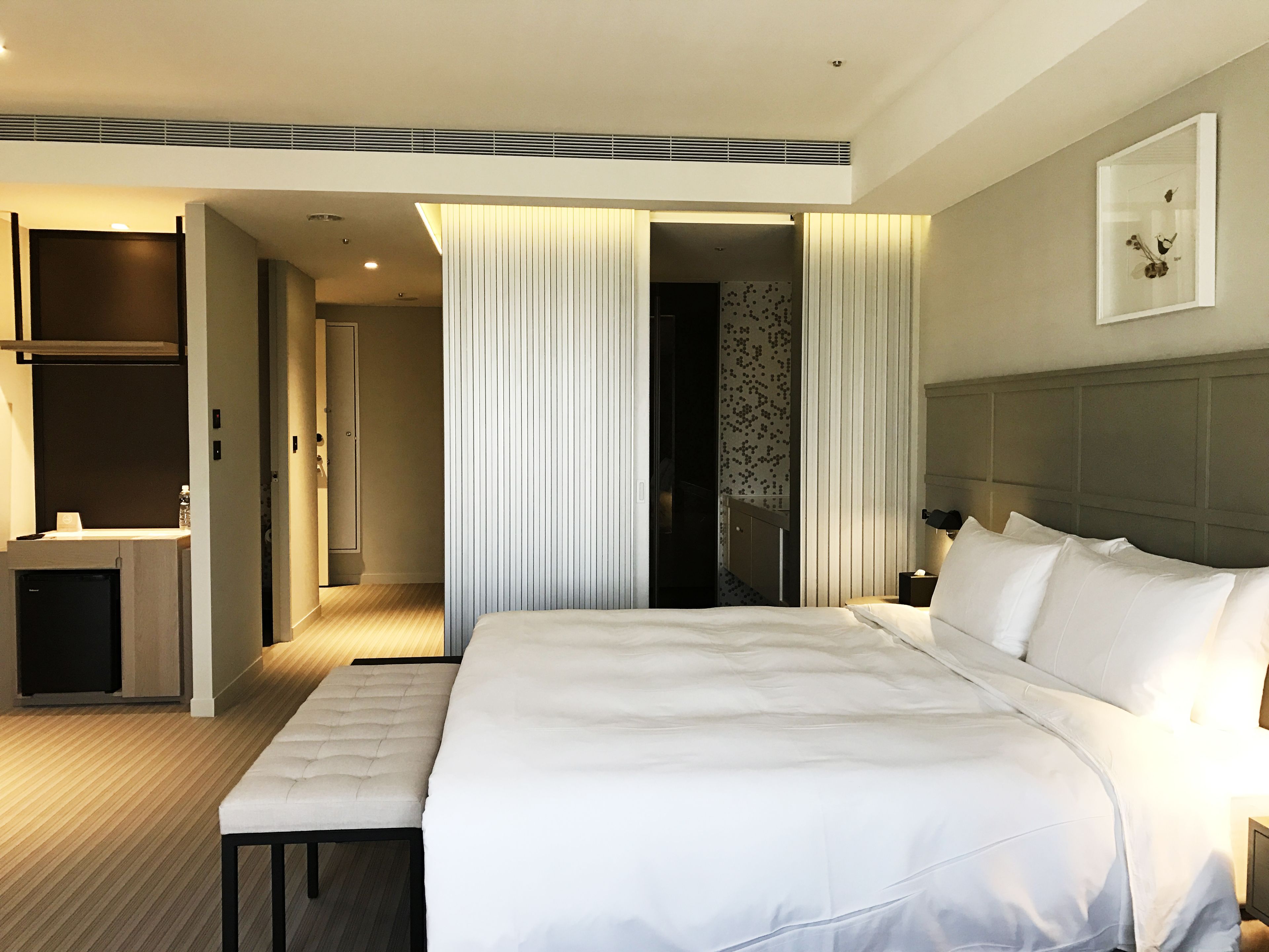 Une chambre d’hôtel moderne avec un grand lit, une table de chevet, un banc, une armoire et une salle de bain visible à travers une porte ouverte.