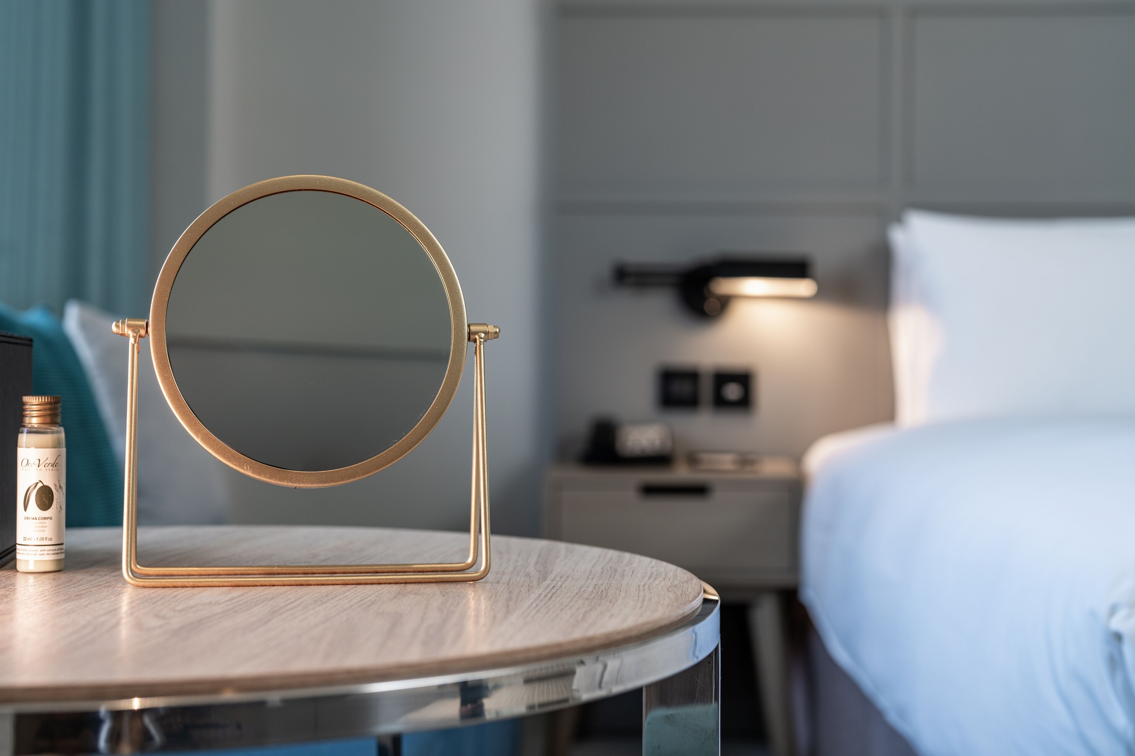 Un miroir rond avec un cadre doré est posé sur une table ronde. Un flacon de lotion se trouve à côté du miroir. Un lit aux draps blancs est visible à l’arrière-plan.