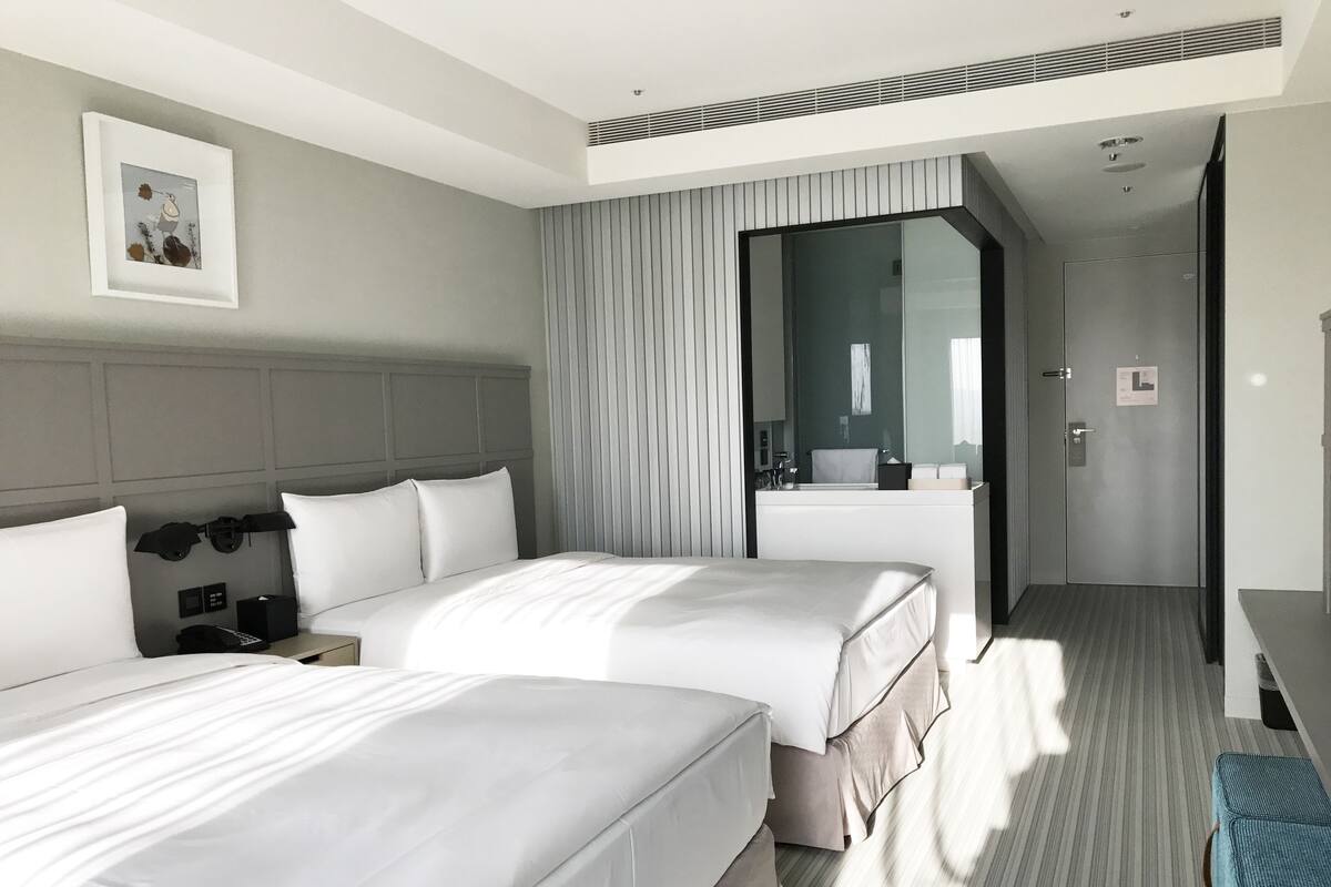 A modern hotel room with two beds, a large mirror, a desk, and a bathroom visible through a glass door.