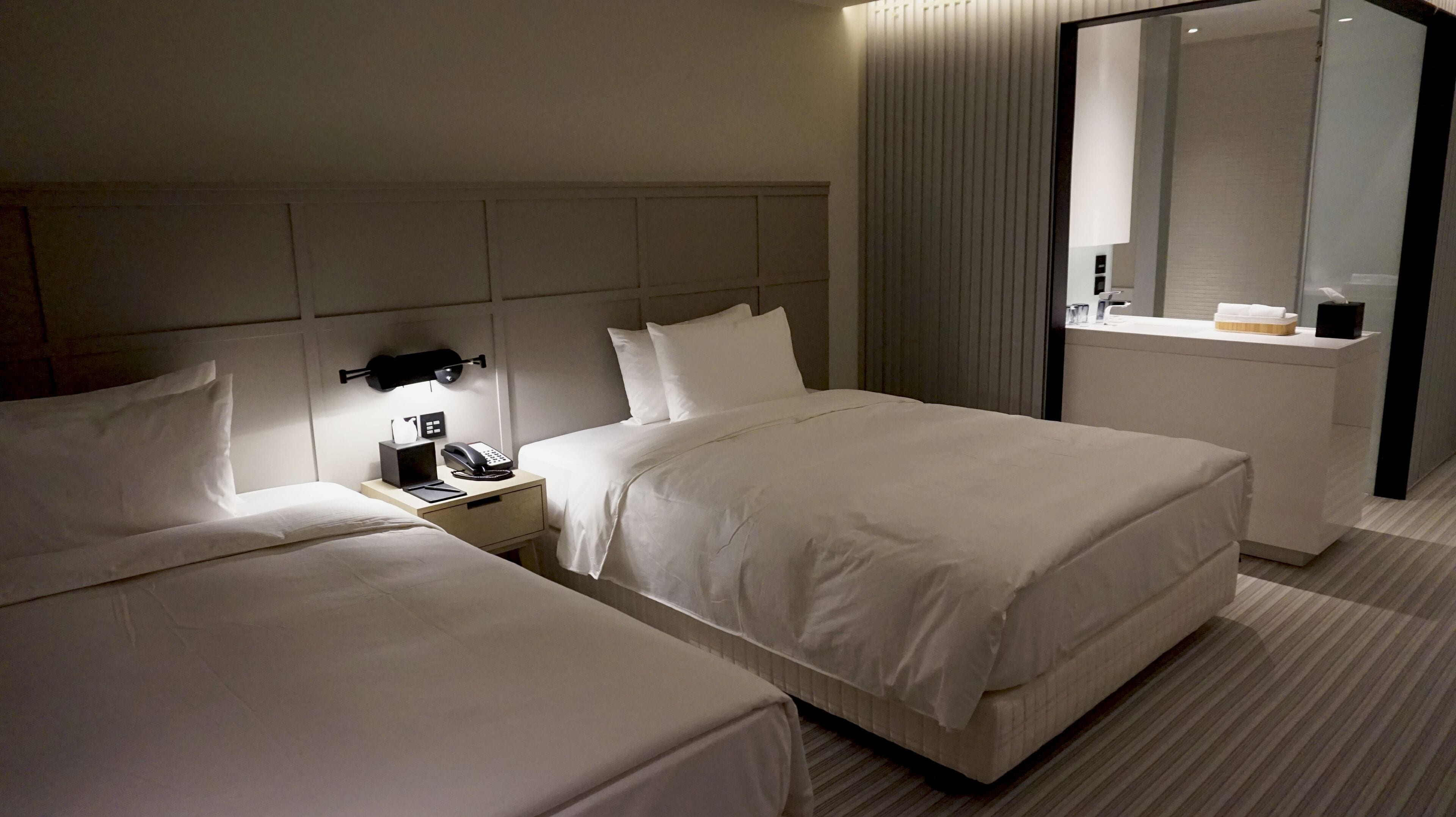 A hotel room with two beds, a bedside table with a phone and a lamp, a small desk with a mirror, and a bathroom visible through a glass partition.
