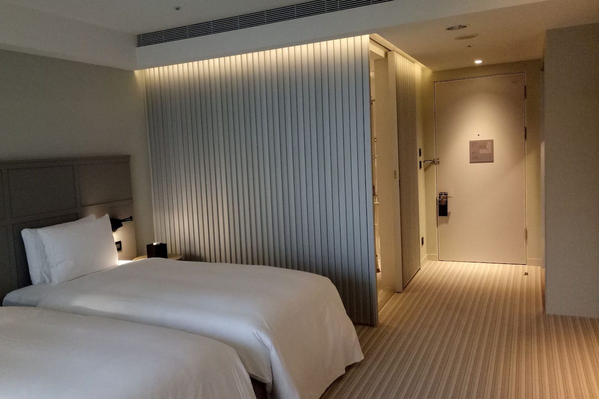 A hotel room with two beds, a headboard, a wall-mounted clock, and a door leading to another room.