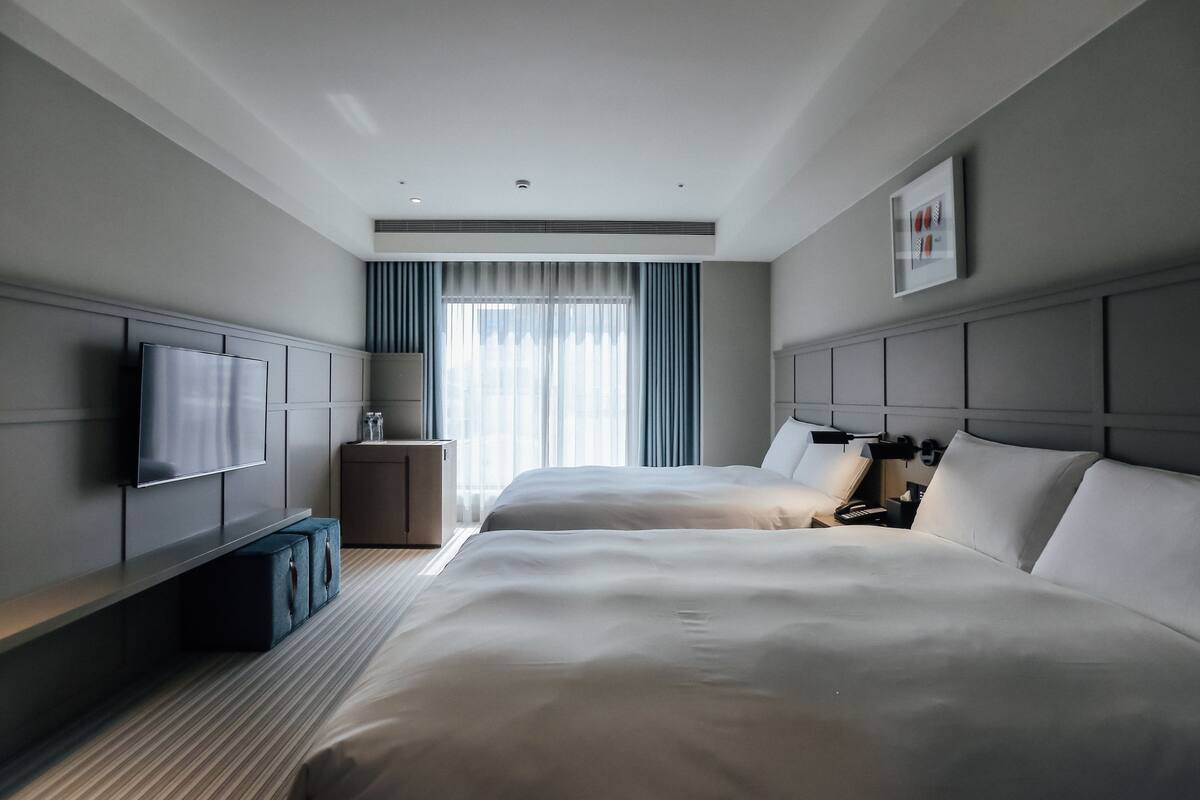 A hotel room with two beds, a TV, a bedside table, a lamp, and a window with curtains.