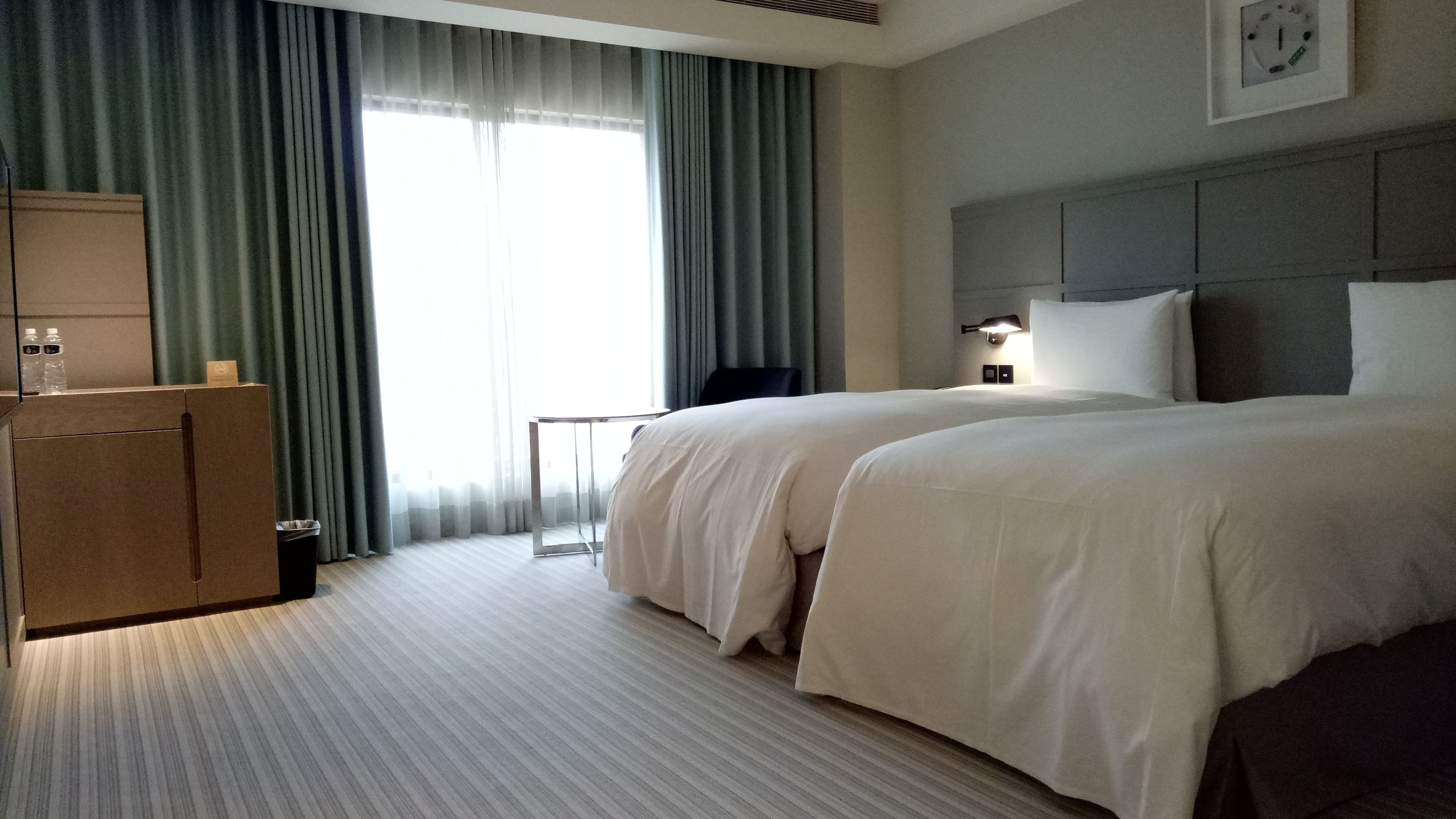 A hotel room with two beds, a desk, a chair, a clock on the wall, and a window with curtains.