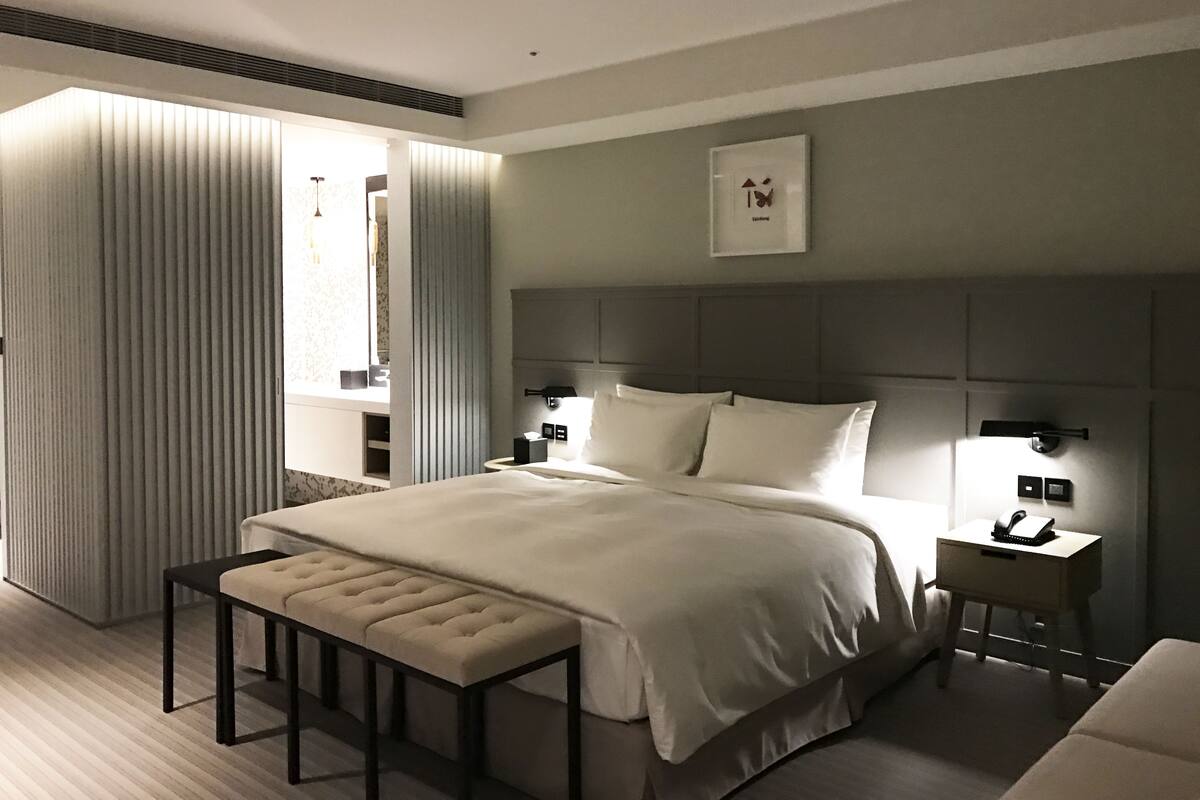 A modern hotel room with a large bed, a bedside table, a nightstand, a bench, and a window with blinds.