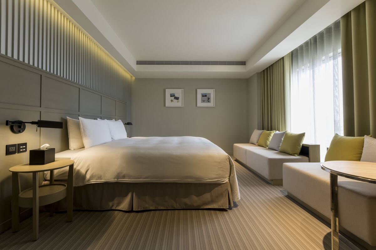 A modern hotel room with a large bed, a sofa, a small table, and a bedside table with a lamp.