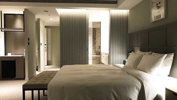 A modern hotel room with a large bed, a bedside table, a bench, a wardrobe, and a bathroom visible through a sliding door. - Greet Inn (Kaohsiung)