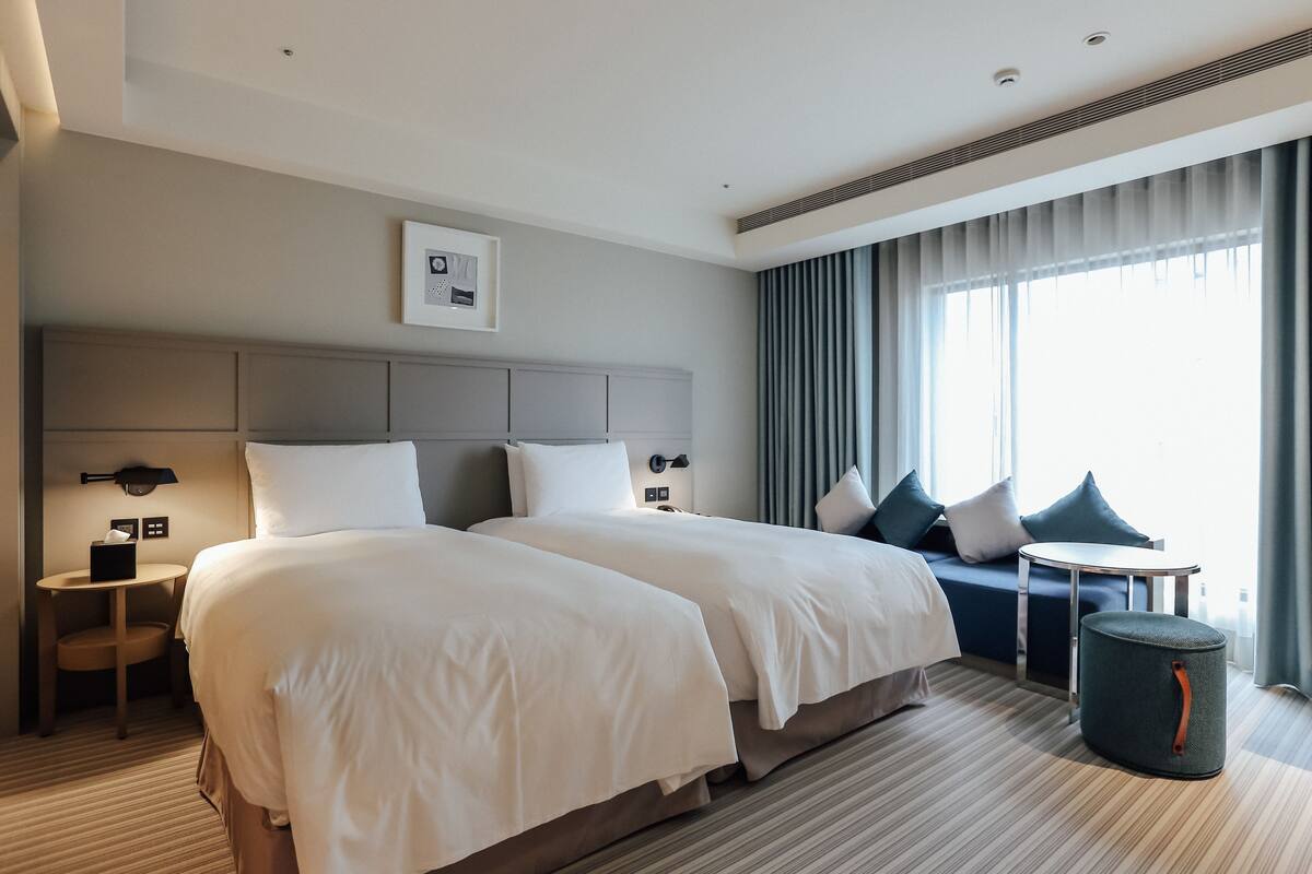 A hotel room with two beds, a round table, a small stool, and a large window with curtains.