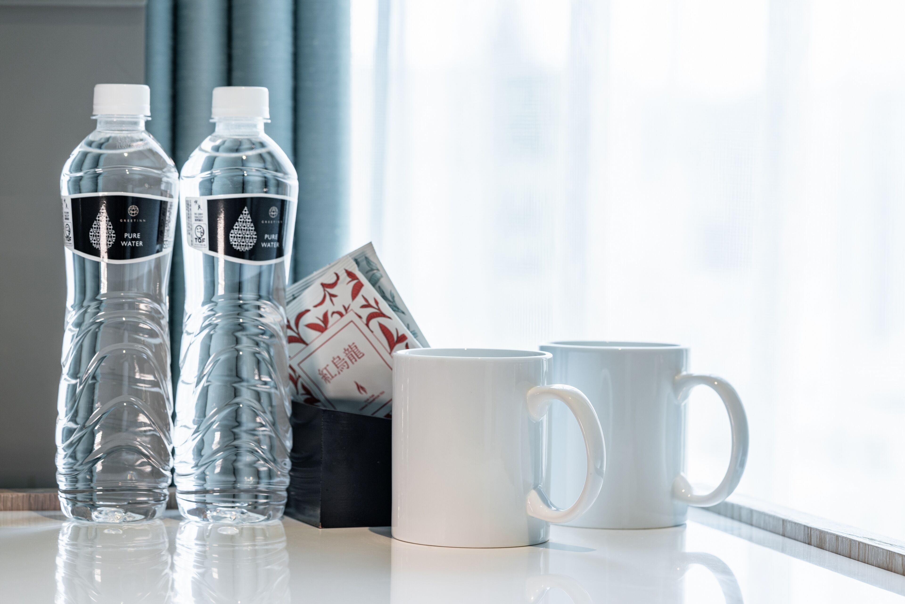 Deux bouteilles d’eau transparente et une tasse blanche sont posées sur une surface réfléchissante, près d’une fenêtre ornée de rideaux.