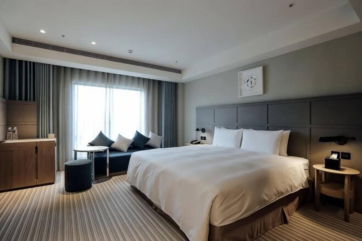 A hotel room with a large bed, a desk, a chair, a small table, a wardrobe, and a window with curtains.
