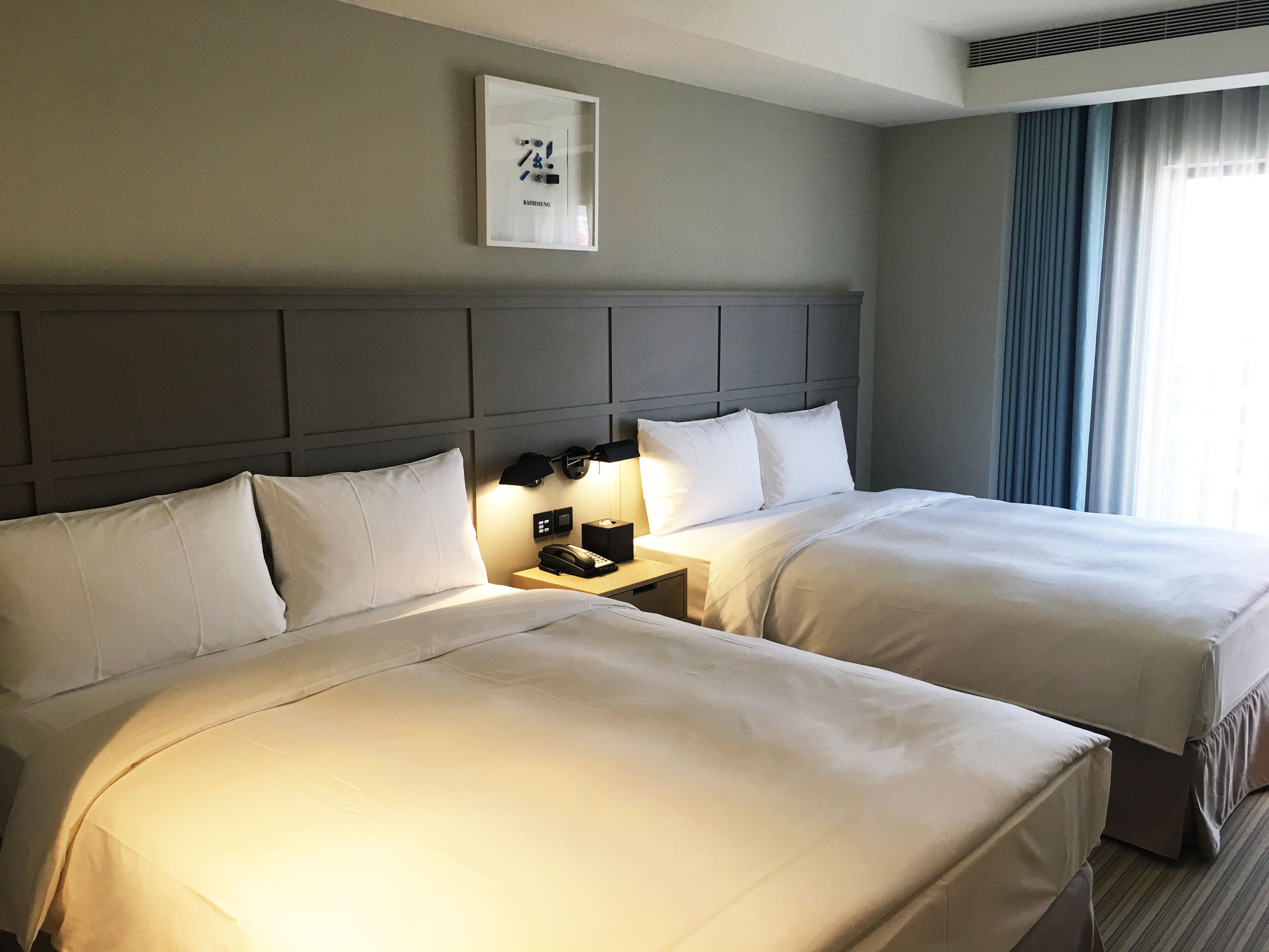 A hotel room with two beds, a headboard, bedside lamps, a telephone, and a framed picture on the wall.