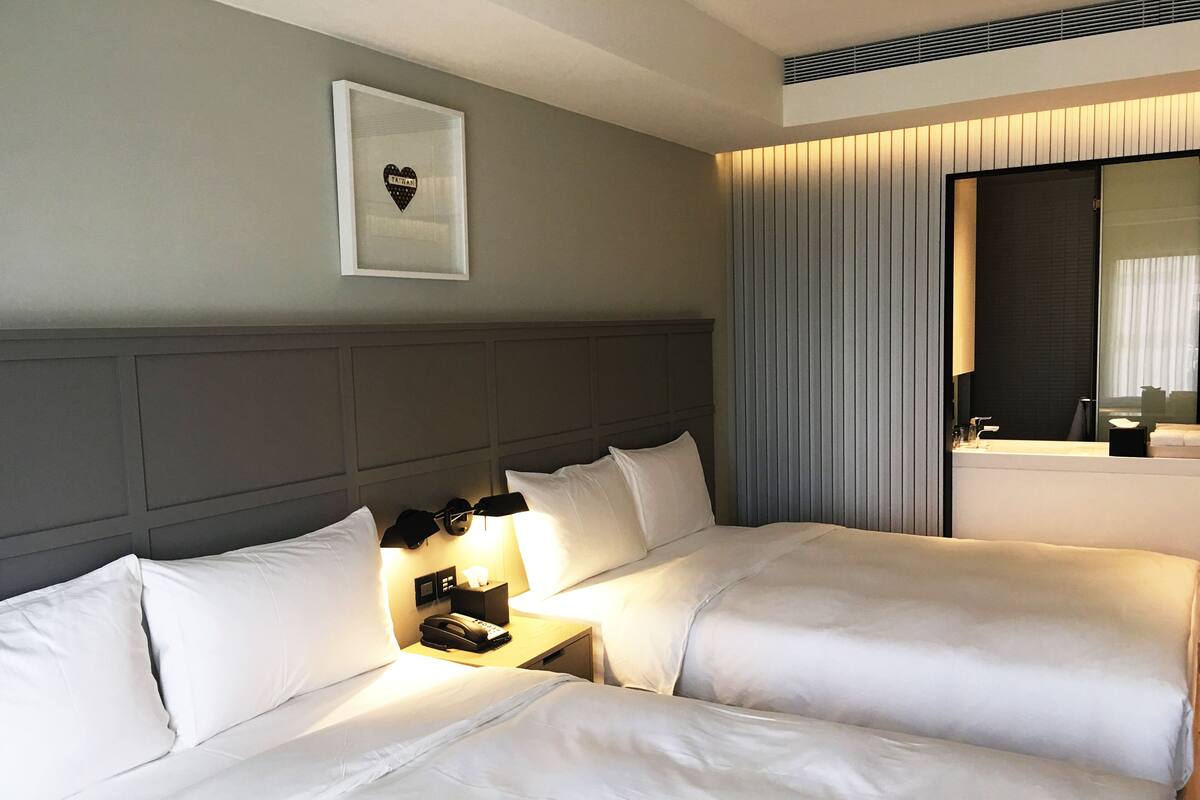 A hotel room with two beds, a bedside table with a lamp, a framed picture on the wall, and a large mirror.