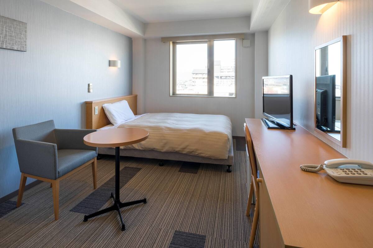 Quarto de hotel com cama, escrivaninha, telefone, cadeira, televisão e janela com vista para a cidade.
