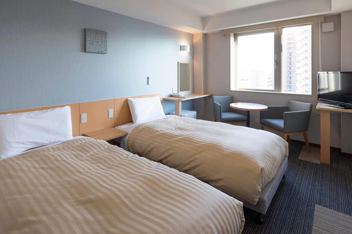 Quarto de hotel com duas camas, uma escrivaninha com televisão, uma mesinha e uma cadeira.