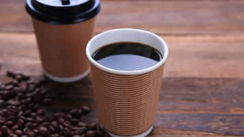 Dos vasos de café de cartón con tapas sobre una superficie de madera, rodeados de granos de café.