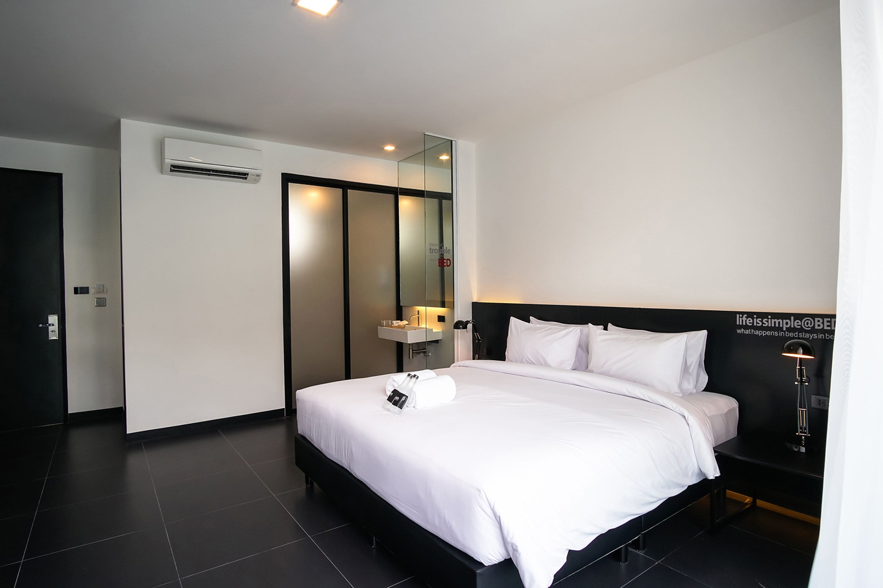 A modern hotel room with a large bed, a bedside table, a mirror, and an air conditioning unit.