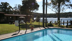 A swimming pool with a wooden deck, surrounded by a fence and potted plants. - Lake Edge Holiday Units (Burrill Lake)