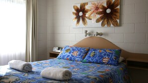 A hotel room with a bed featuring a blue bedspread with marine life patterns, two pillows with similar designs, a wall-mounted radio, and a large floral artwork above the bed. - Lake Edge Holiday Units (Burrill Lake)