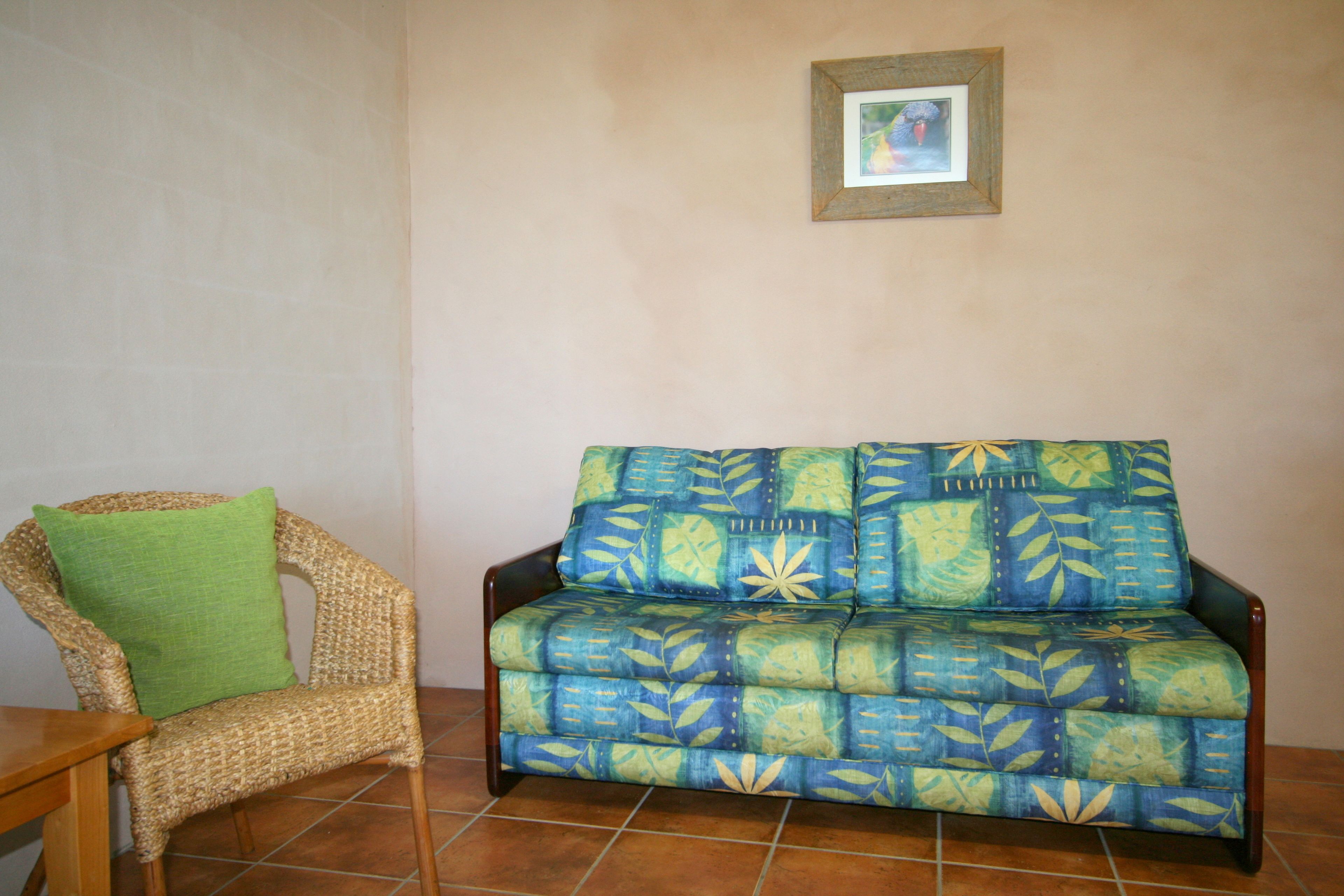 A wicker chair with a green cushion and a patterned sofa in a room with tiled flooring.