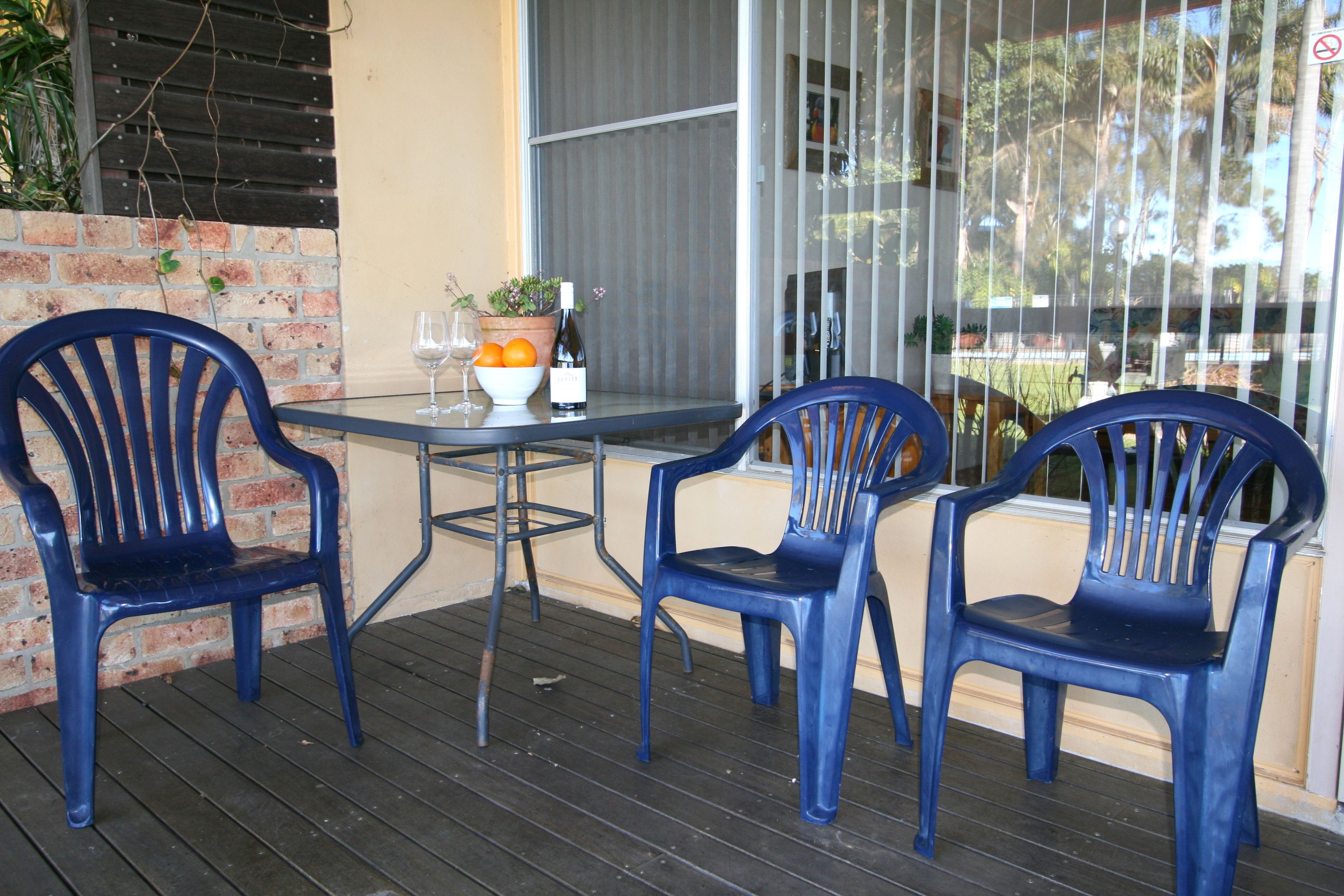 A table with two blue chairs, a bowl of fruit, a wine glass, and a bottle of wine on a balcony with a brick wall and a window.