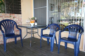 A table with two blue chairs, a bowl of fruit, a wine glass, and a bottle of wine on a balcony with a brick wall and a window. - Lake Edge Holiday Units (Burrill Lake)