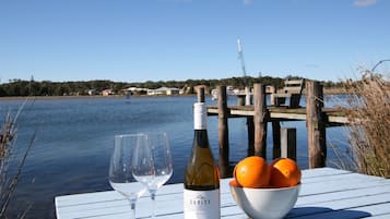 A table with a wine bottle and a bowl of oranges by a lake with a dock and houses in the background.