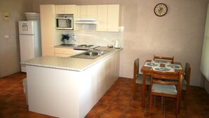 A compact kitchen with a central island, a microwave, a stove, and a sink. There is a dining table with chairs and a clock on the wall. - Lake Edge Holiday Units (Burrill Lake)