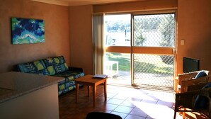 A living room with a sofa, a table, and a TV. There is a large window with a view of the outdoors. - Lake Edge Holiday Units (Burrill Lake)