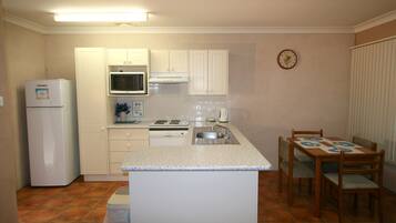 Een kleine keuken met witte kastjes, een magnetron, een fornuis en een spoelbak. Er staat een eettafel met stoelen en een klok hangt aan de muur.