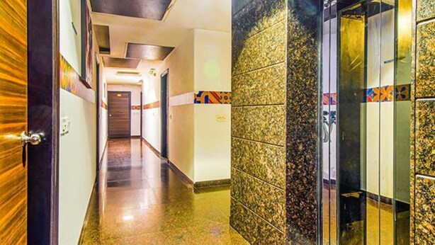 A hotel corridor with a wooden door on the left, a tiled wall with a geometric pattern on the right, and a glass elevator door at the end.