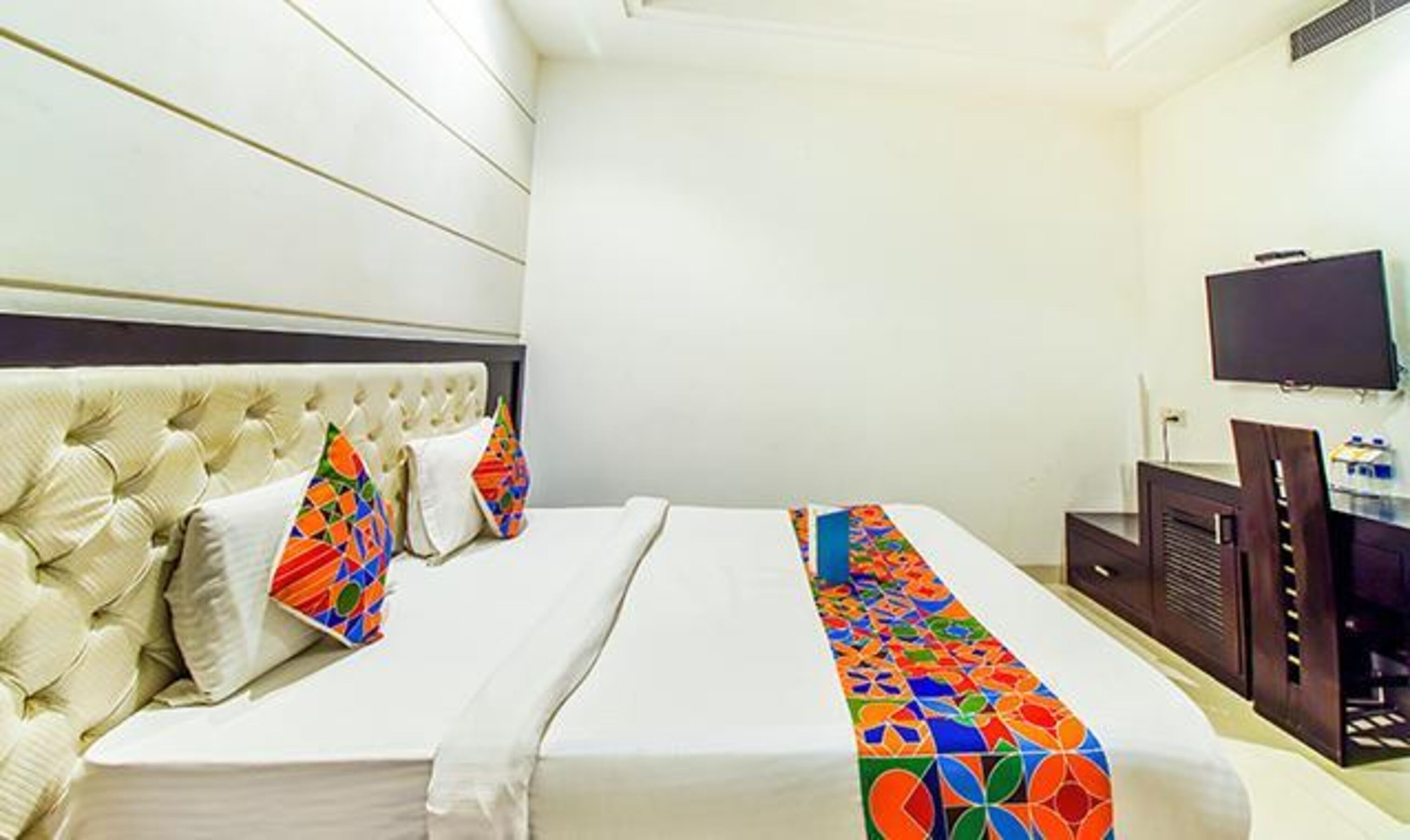 A hotel room with a tufted headboard, a bed with colorful pillows, a TV on the wall, and a desk with a chair.