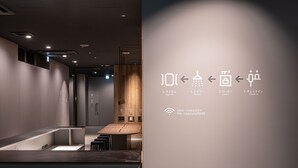 A modern hotel lobby with a reception desk, a wooden reception counter, and a wall with directional signs and Wi-Fi details. - Capsule Plus Yokohama Sauna & Capsule (Yokohama)