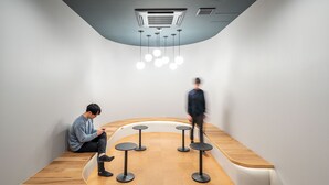 A modern interior with a curved wooden bench seating area, a ceiling-mounted air conditioning unit, and a row of hanging spherical lights. - Capsule Plus Yokohama Sauna & Capsule (Yokohama)