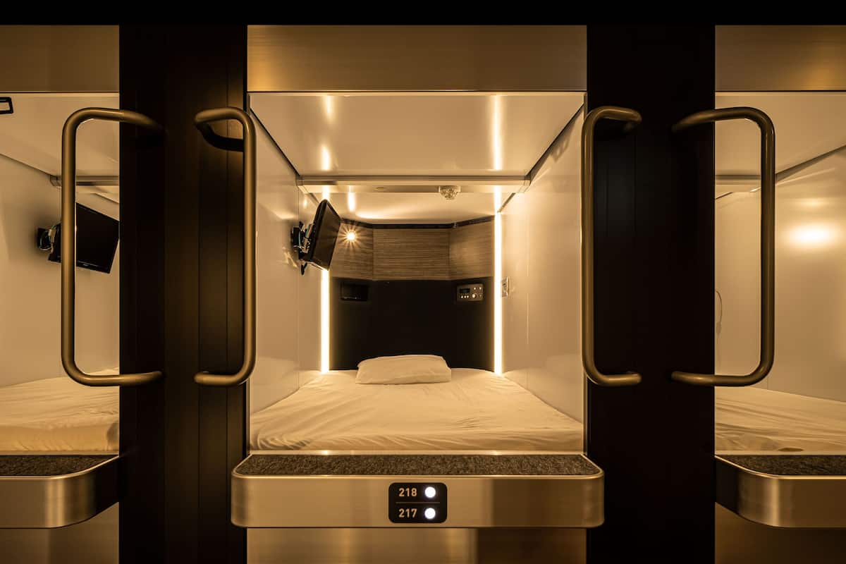 A compact, modern train cabin with a single bed, a small television, and a control panel with room numbers.