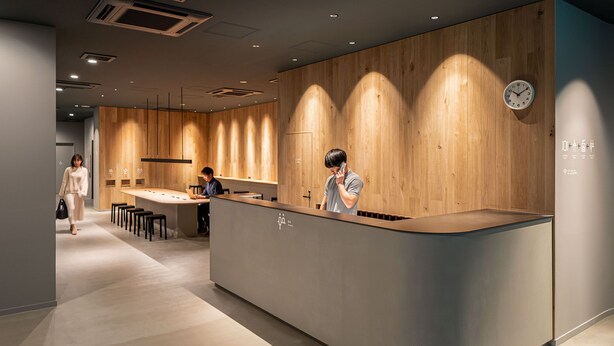 A modern reception area with a wooden reception desk, a clock on the wall, and a seating area with chairs. - Capsule Plus Yokohama Sauna & Capsule (Yokohama)