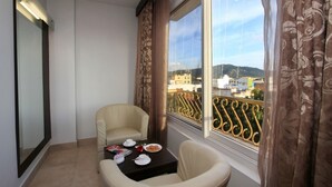 A balcony with a view of buildings and hills, a chair, a small table with a plate of food, and curtains.