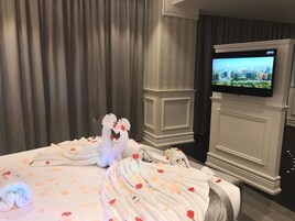 Habitación de hotel con una cama decorada con pétalos de rosa y toallas con forma de cisne, un televisor montado en la pared y vistas a la ciudad.