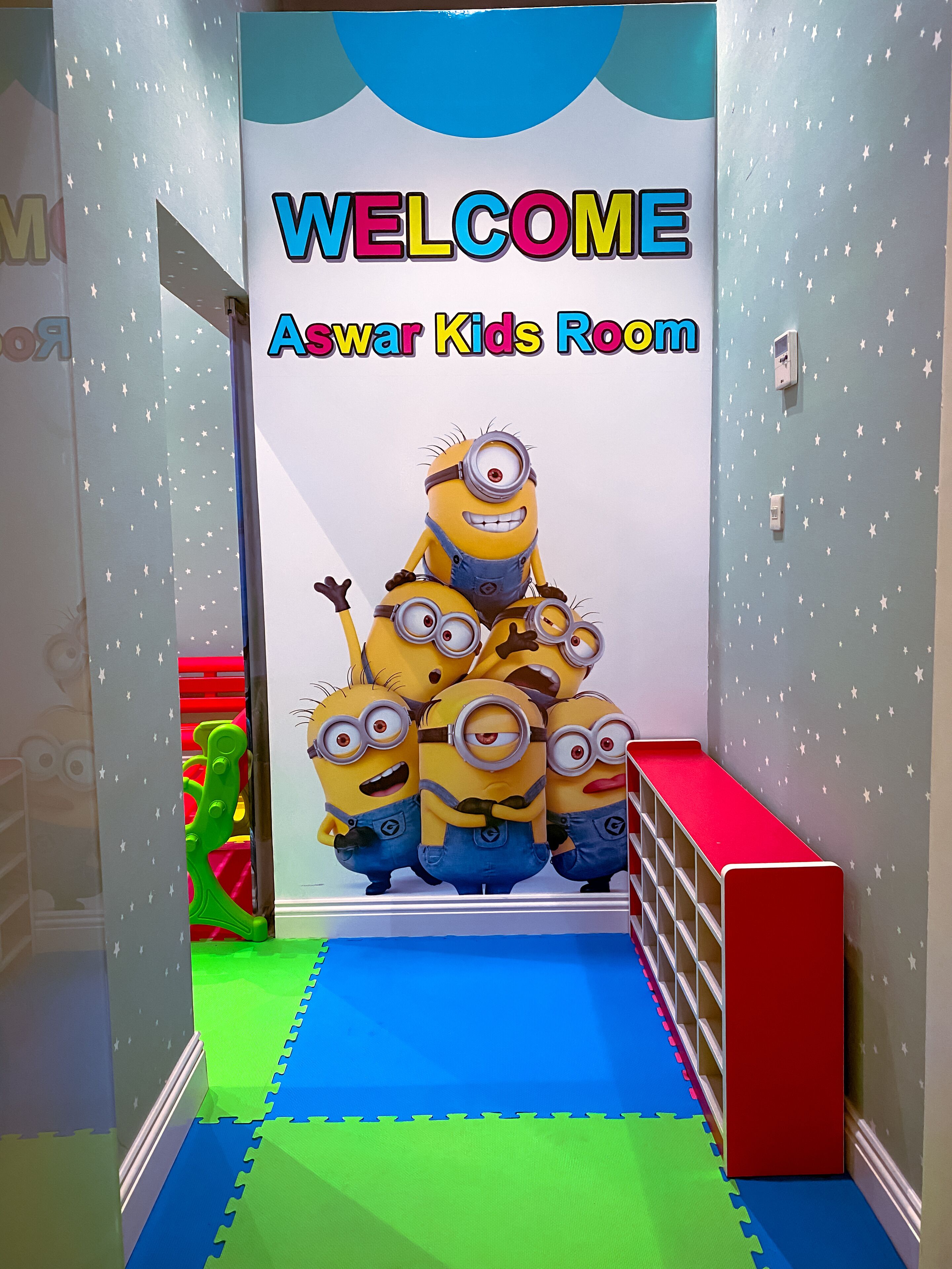 A children's play area with a 'Welcome Aswar Kids Room' sign, Minion characters, and colorful play mats.